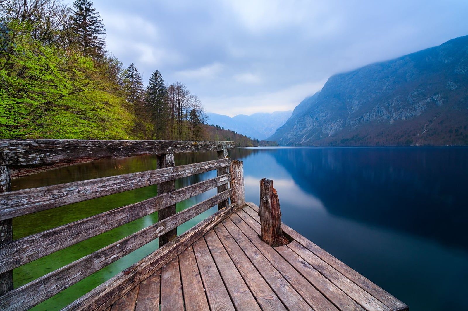 Ein Holzsteg blickt auf das ruhige blaue Wasser des Bohinj-Sees, umrahmt von bewaldeten Ufern und den Julischen Alpen im Triglav-Nationalpark.