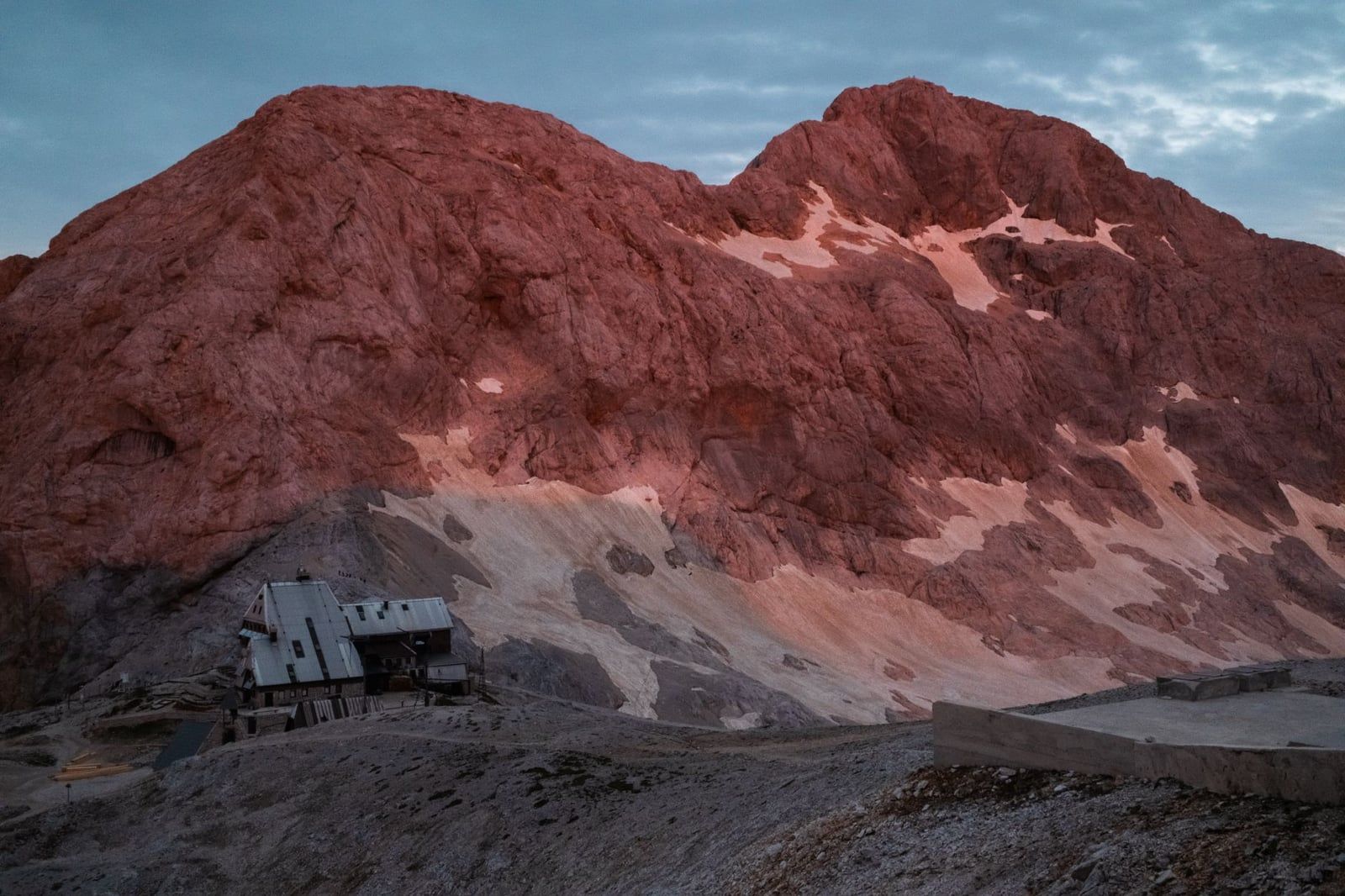 Eine Berghütte liegt auf einem felsigen Grat unter gewaltigen Gipfeln im Morgenlicht im Triglav-Nationalpark.
