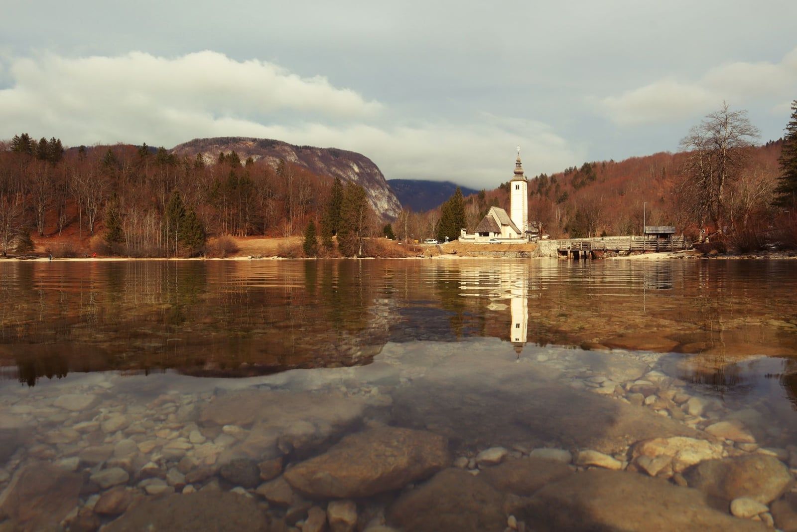 The Church of St. John the Baptist reflected in the clear waters of Lake Bohinj against autumn-colored hills in Triglav National Park.