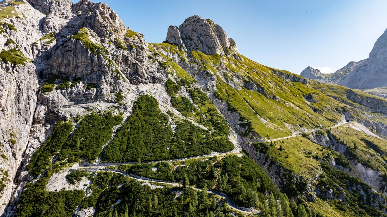 The narrow Mangart road winds uphill through steep slopes and a rock tunnel toward the Mangart saddle in Triglav National Park.